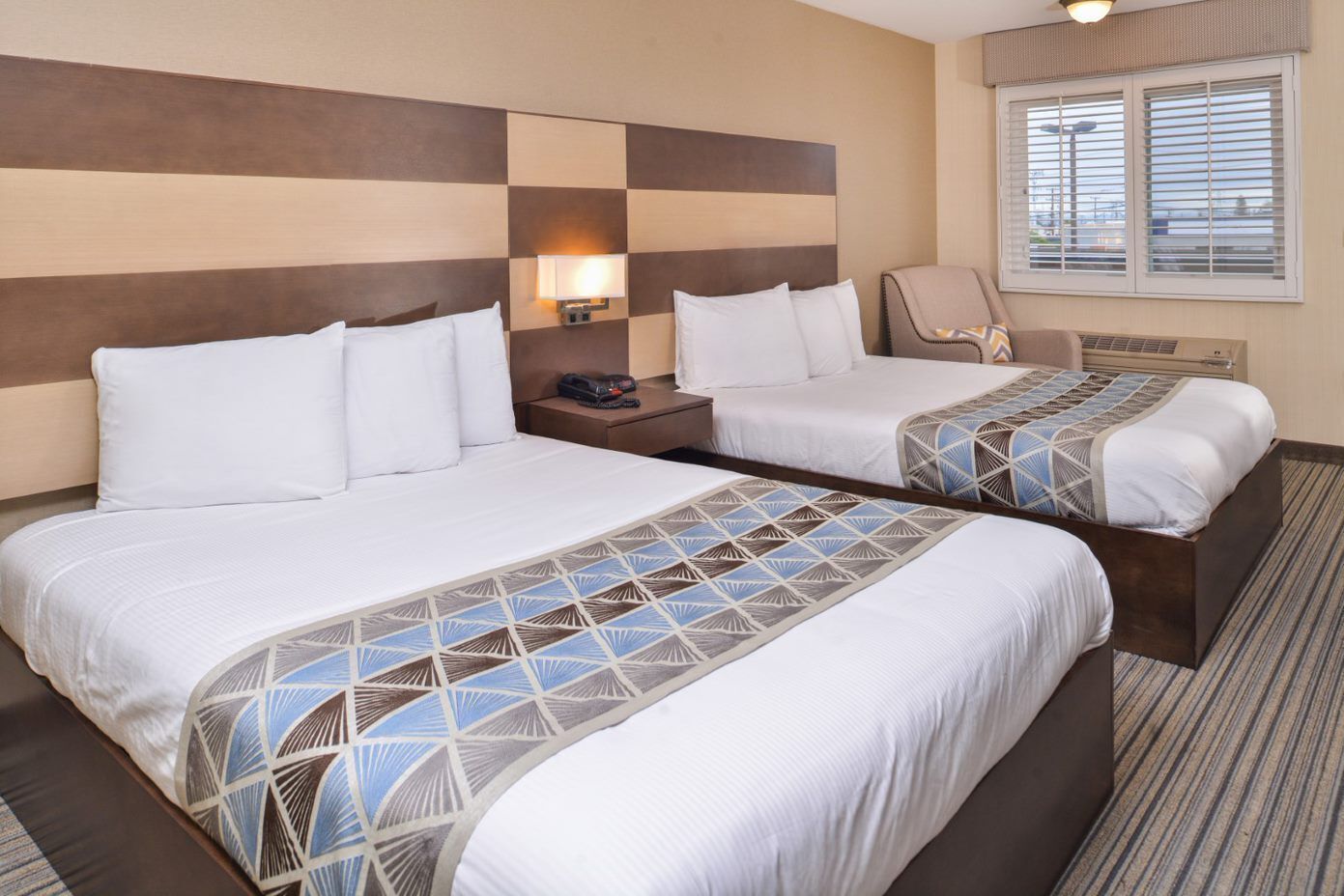 A hotel room with two beds, a nightstand, a lamp, a chair, and a window with blinds.