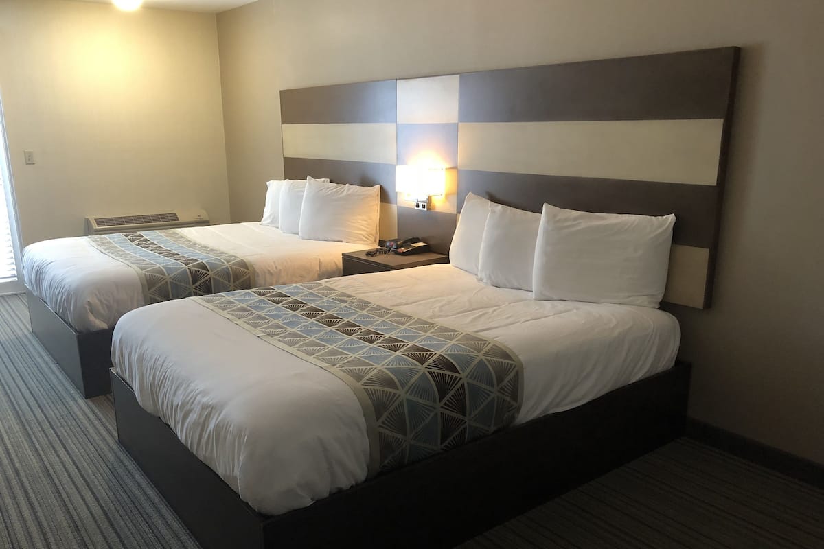 Ein Hotelzimmer mit zwei Betten, einem großen, an der Wand befestigten Kopfteil, einem Nachttisch mit Lampe und einem Teppichboden.