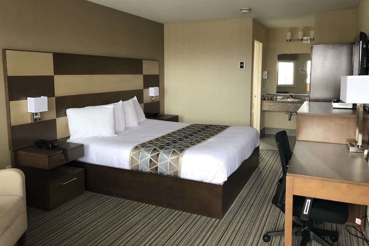Ein Hotelzimmer mit einem großen Bett, Nachttischen, einem Schreibtisch, einem Sessel und einem Badezimmer mit Waschbecken und Spiegel. Ein Telefon ist ebenfalls vorhanden.