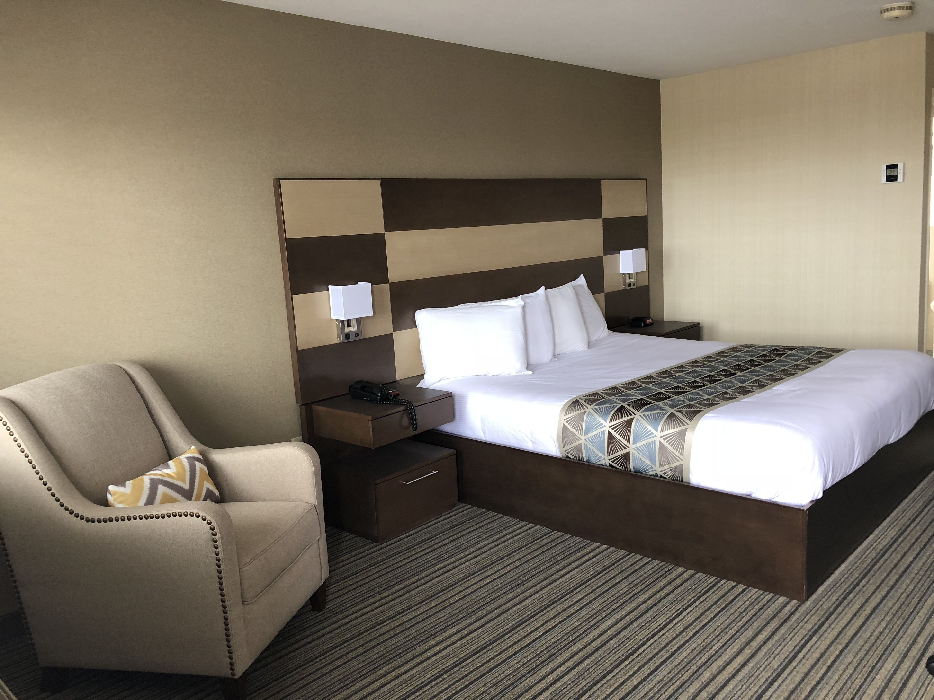 A hotel room with a large bed, a bedside table with a lamp, a chair, and a carpeted floor.