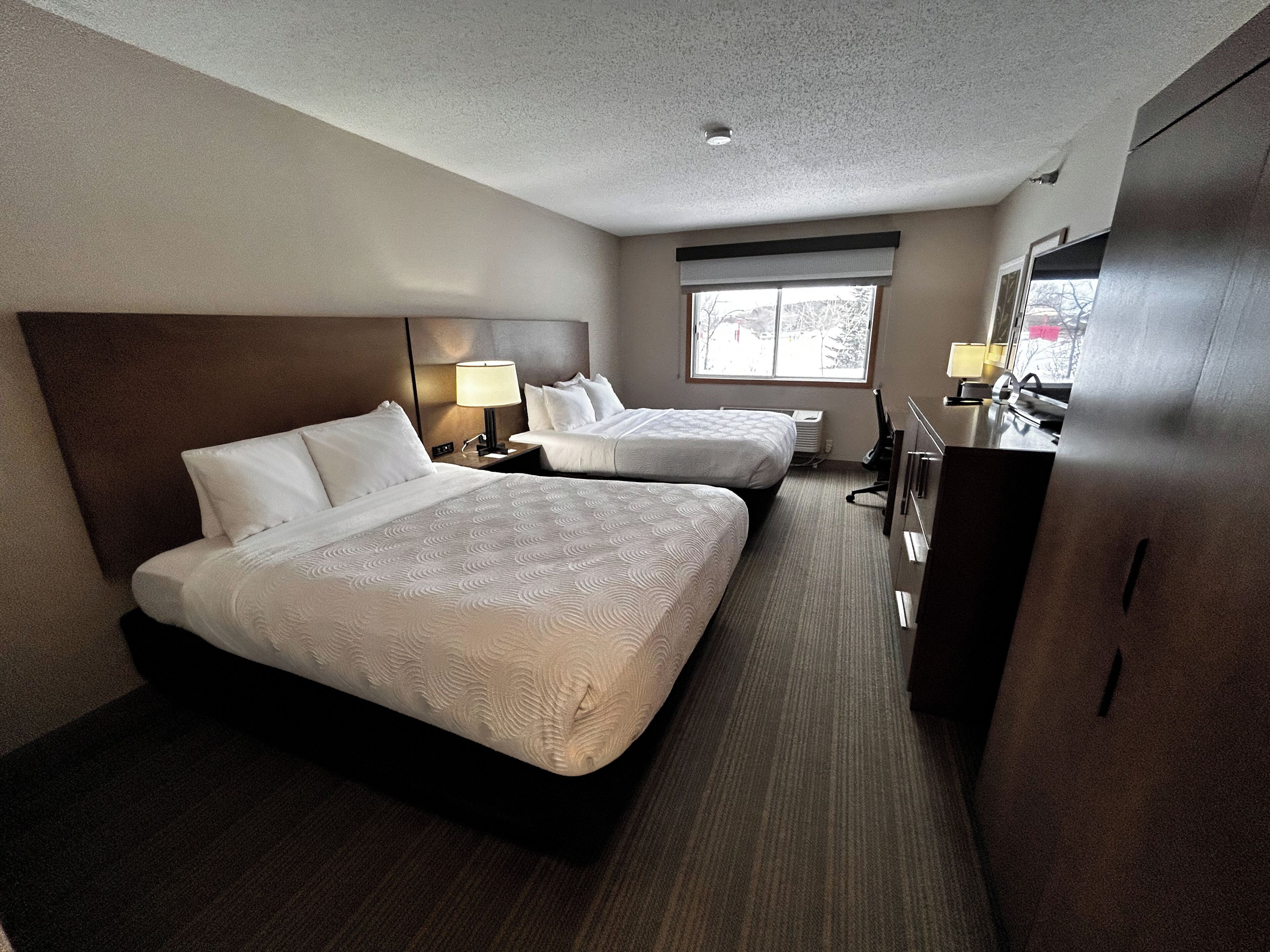 A hotel room with two beds, a desk, a chair, a lamp, and a window.