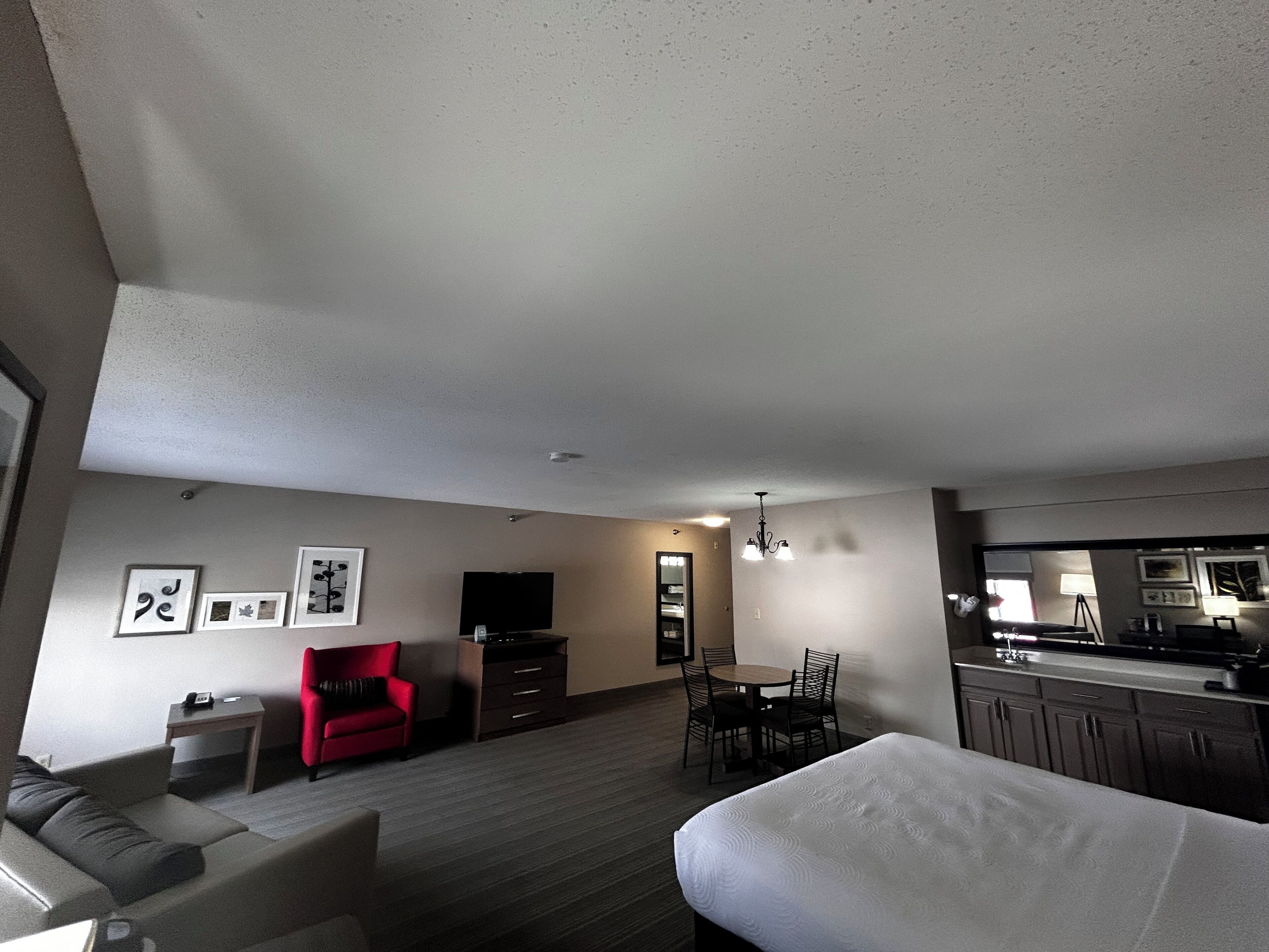 A modern hotel room with a sofa, a red chair, a dining table with chairs, a TV, a kitchen area, and a large bed.