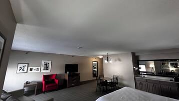 A modern hotel room with a sofa, a red chair, a dining table with chairs, a TV, a kitchen area, and a large bed.