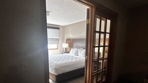 A hotel room with a bed, a nightstand, a lamp, and a view through a door. - Country Inn & Suites by Radisson, Baxter, MN (Brainerd)
