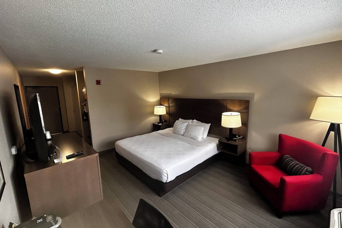 A hotel room with a bed, two bedside tables with lamps, a red armchair, a desk with a computer, and a television.