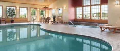 An indoor swimming pool with lounge chairs and tables, large windows, and a wooden ceiling.