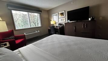 A hotel room with a large bed, a desk, a chair, a television, and a window with a view of trees.