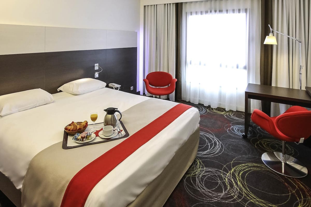 A hotel room with a neatly made bed, breakfast tray, red chairs, and a view of the outdoors through the window.