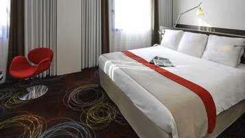 A hotel room with a bed, a red chair, a lamp, and a window with curtains.