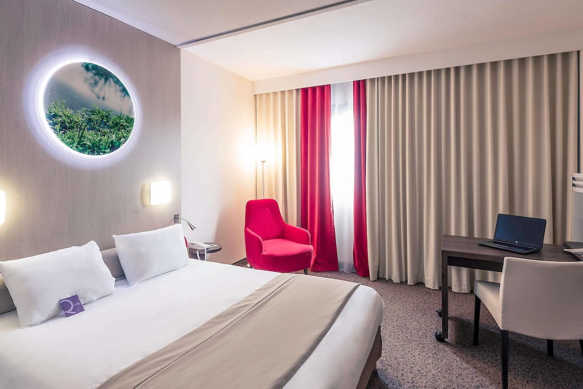 A hotel room with a bed, a desk with a laptop, a red chair, and a round mirror on the wall.