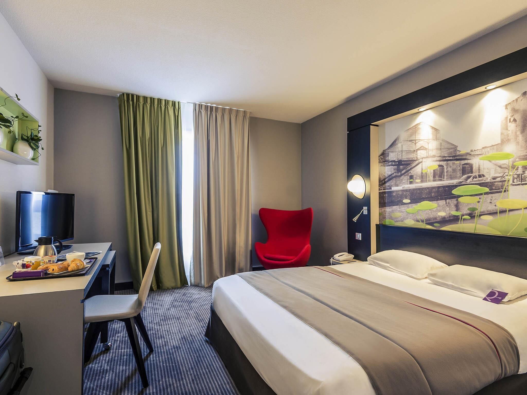 A hotel room with a bed, a desk with a computer, a chair, a painting on the wall, and a window with curtains.