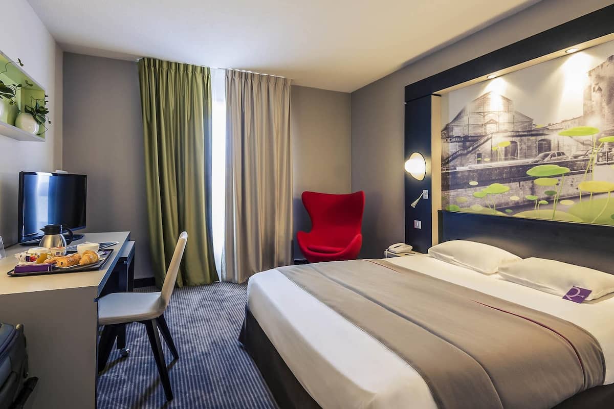A hotel room with a bed, a desk with a computer, a chair, a painting on the wall, and a window with curtains.