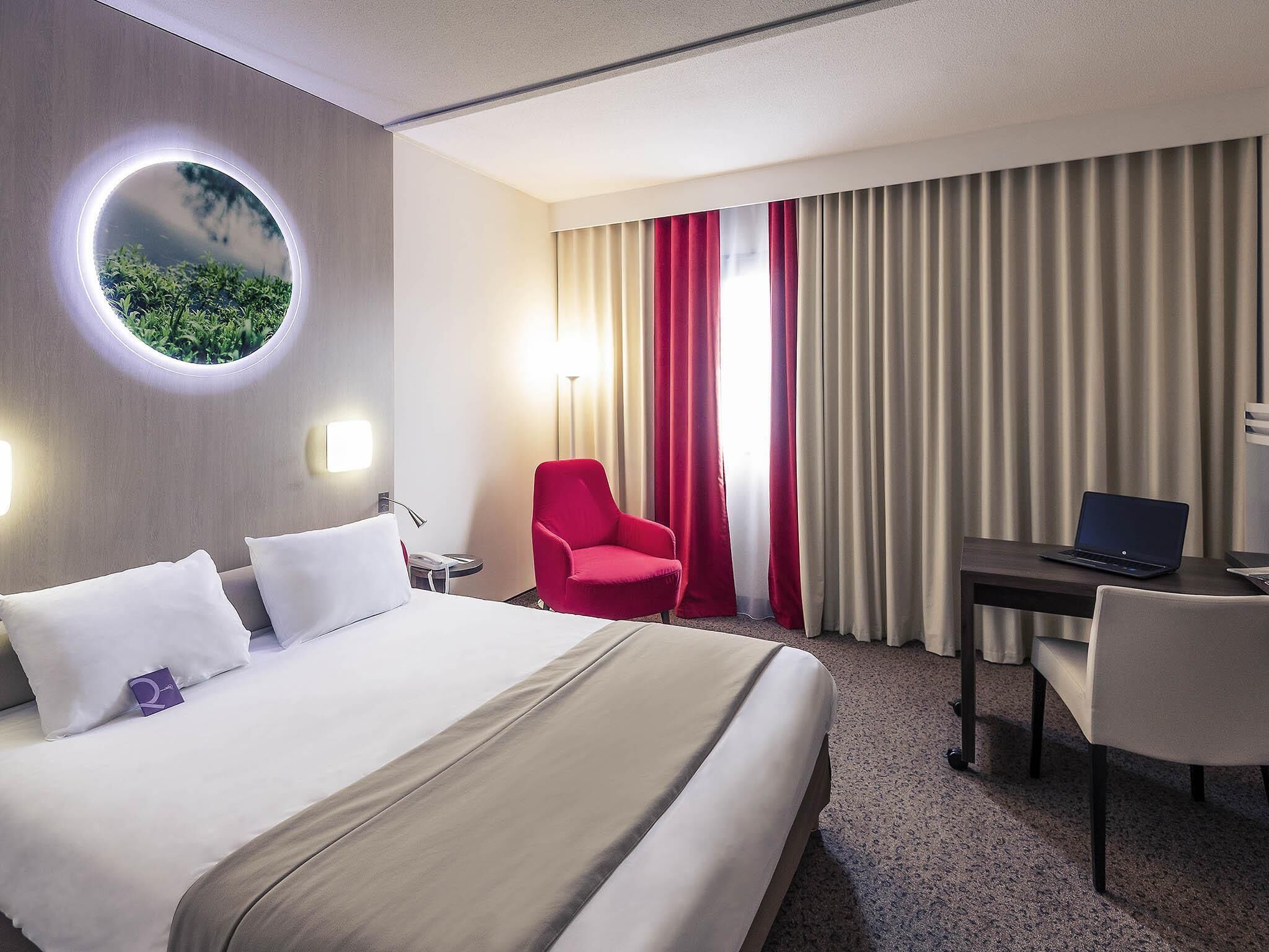 A hotel room with a bed, a desk with a laptop, a red chair, and a round mirror on the wall.
