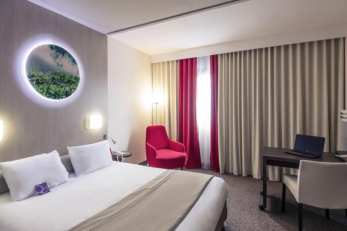 A hotel room with a bed, a desk with a laptop, a red chair, and a round mirror on the wall.