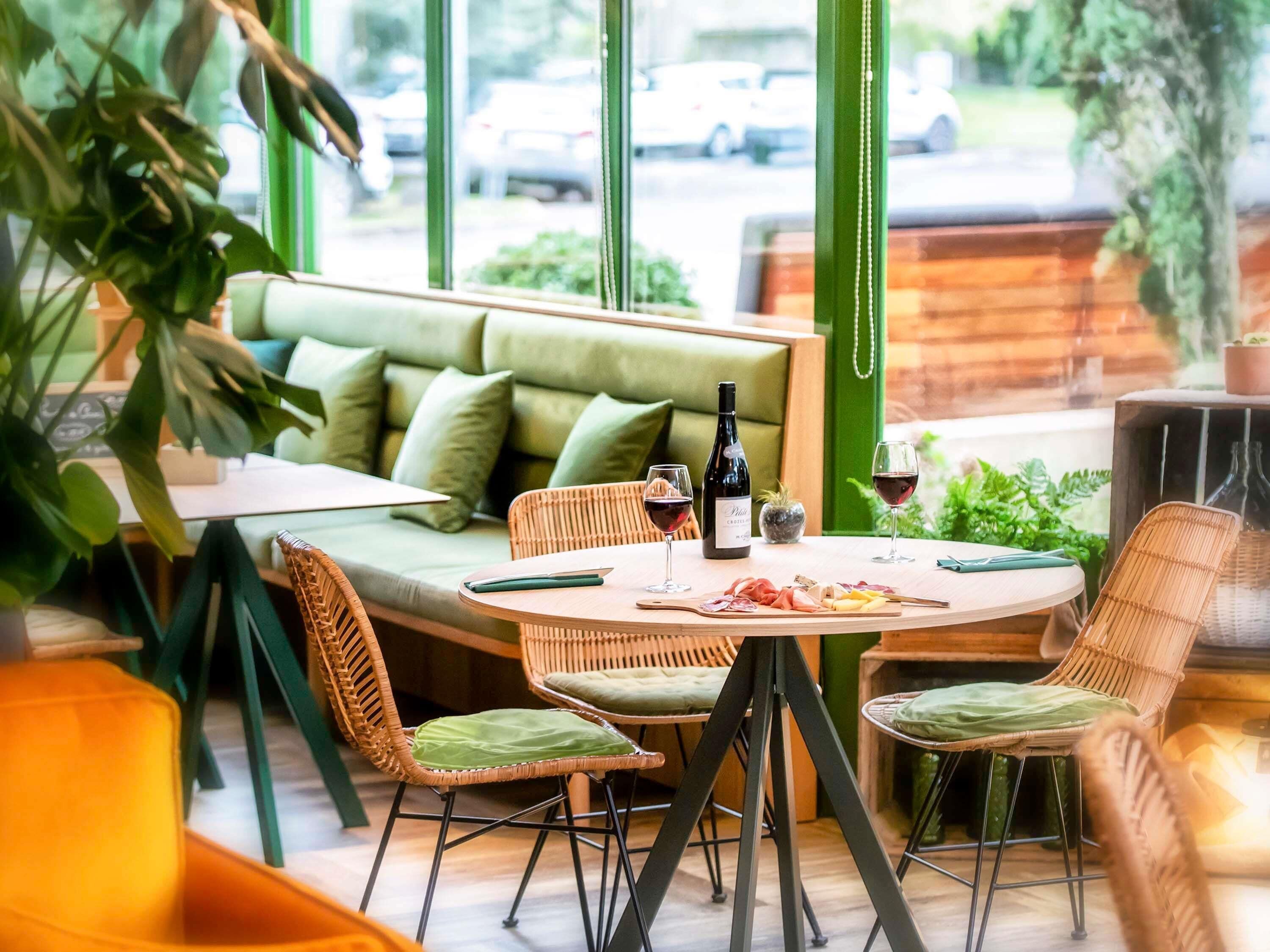 Un coin salon accueillant avec un canapé vert, des fauteuils en osier et une table ronde sur laquelle se trouvent une bouteille et des verres.