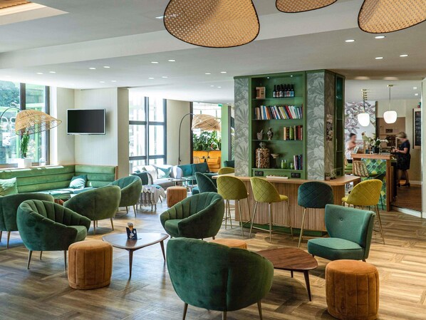Uma área de lounge moderna com poltronas estofadas em verde, uma mesa de centro de madeira e uma estante verde com livros e objetos decorativos.