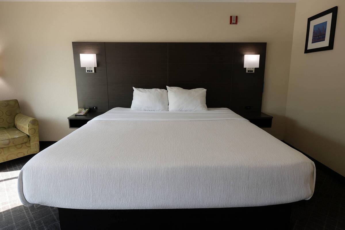 Quarto de hotel com uma cama grande, duas mesas de cabeceira, uma cadeira e luminárias fixadas na parede.