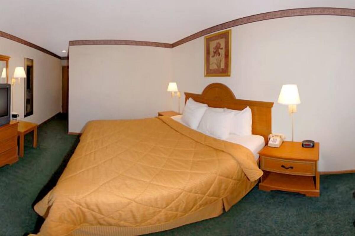 Quarto de hotel com uma cama grande, duas mesas de cabeceira com abajures, uma cadeira vermelha e um quadro na parede.