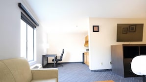 Habitación de hotel moderna con sofá, una pequeña mesa de madera, un escritorio con silla, televisor montado en la pared y ventana con persianas.