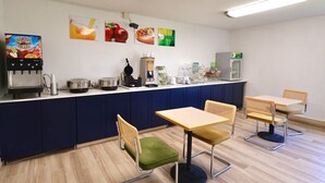 Una kitchenette moderna con cafetera, dispensador de fruta y refrigerador. Hay dos sillas y una mesa en primer plano.