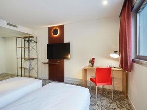 Hotellihuone, jossa on kaksi sänkyä, seinälle asennettu televisio, työpöytä tuolilla ja ikkuna, jossa on verhot.