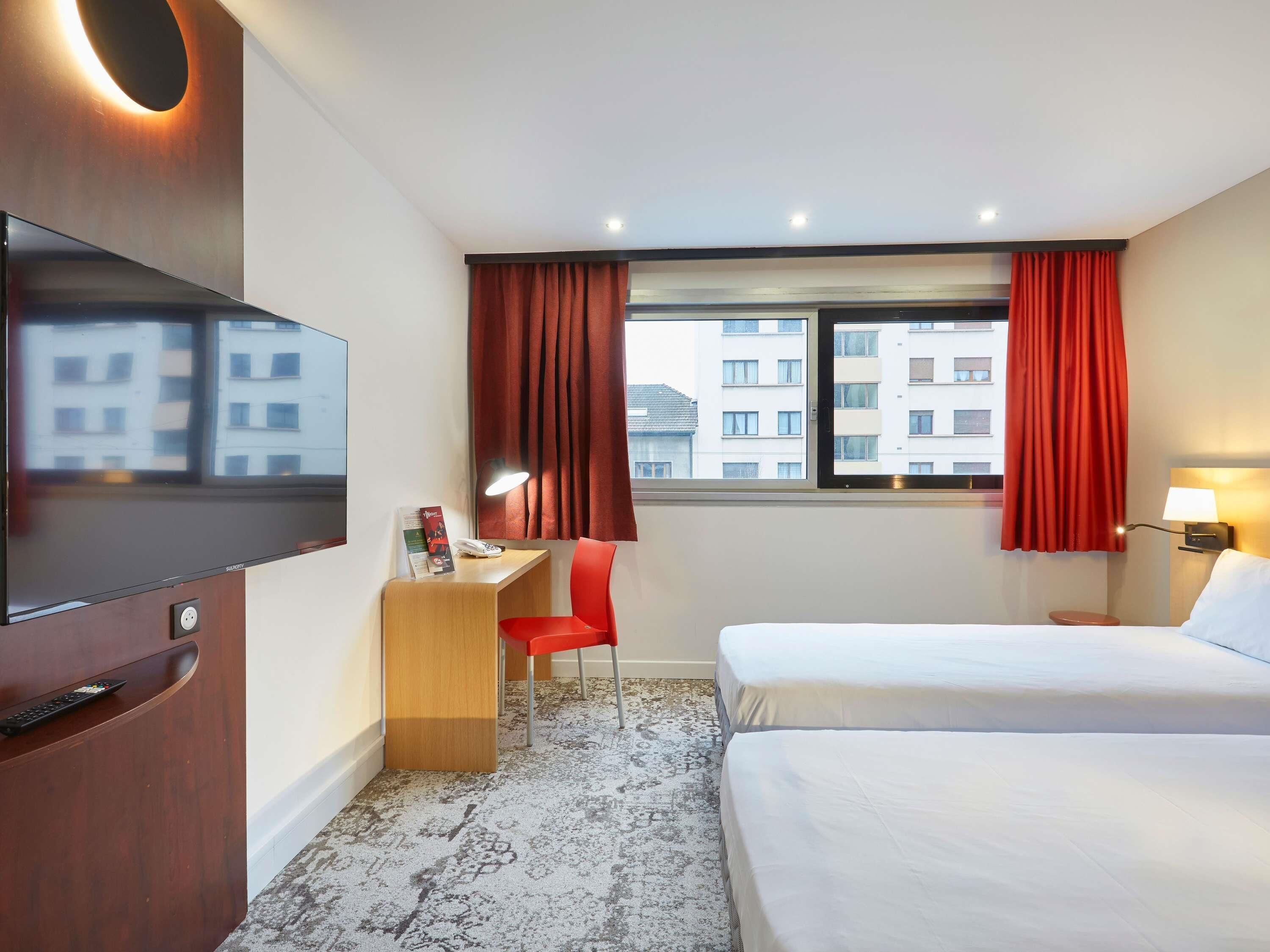 A hotel room with a TV, desk, red chair, two beds, a window with red curtains, and a view of buildings.