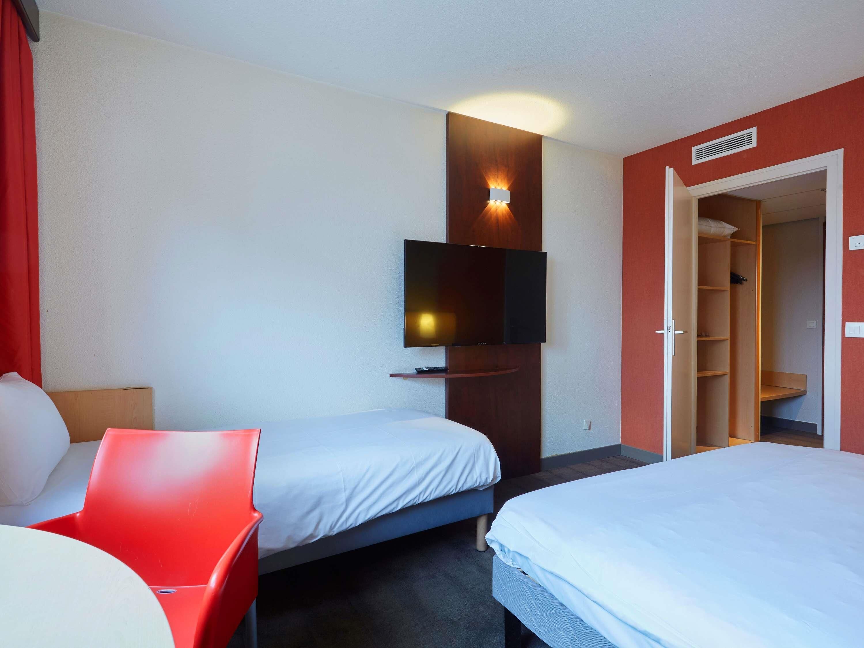 A hotel room with two beds, a TV mounted on the wall, a red chair, and a wardrobe.