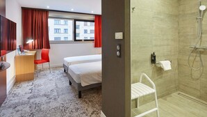 Habitación de hotel con cama, escritorio con silla, zona de ducha y vistas a edificios a través del ventanal.