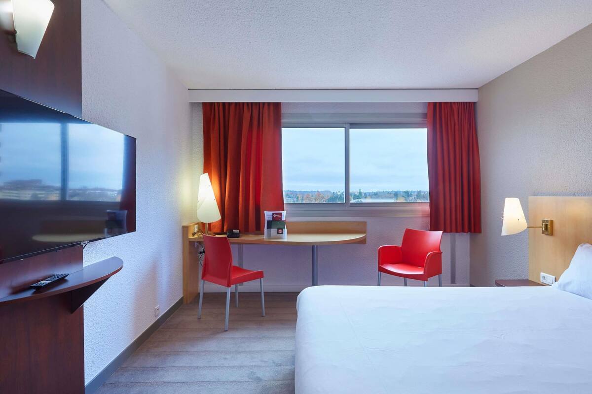 A hotel room with a bed, a desk with a chair, a television, and a window with curtains.