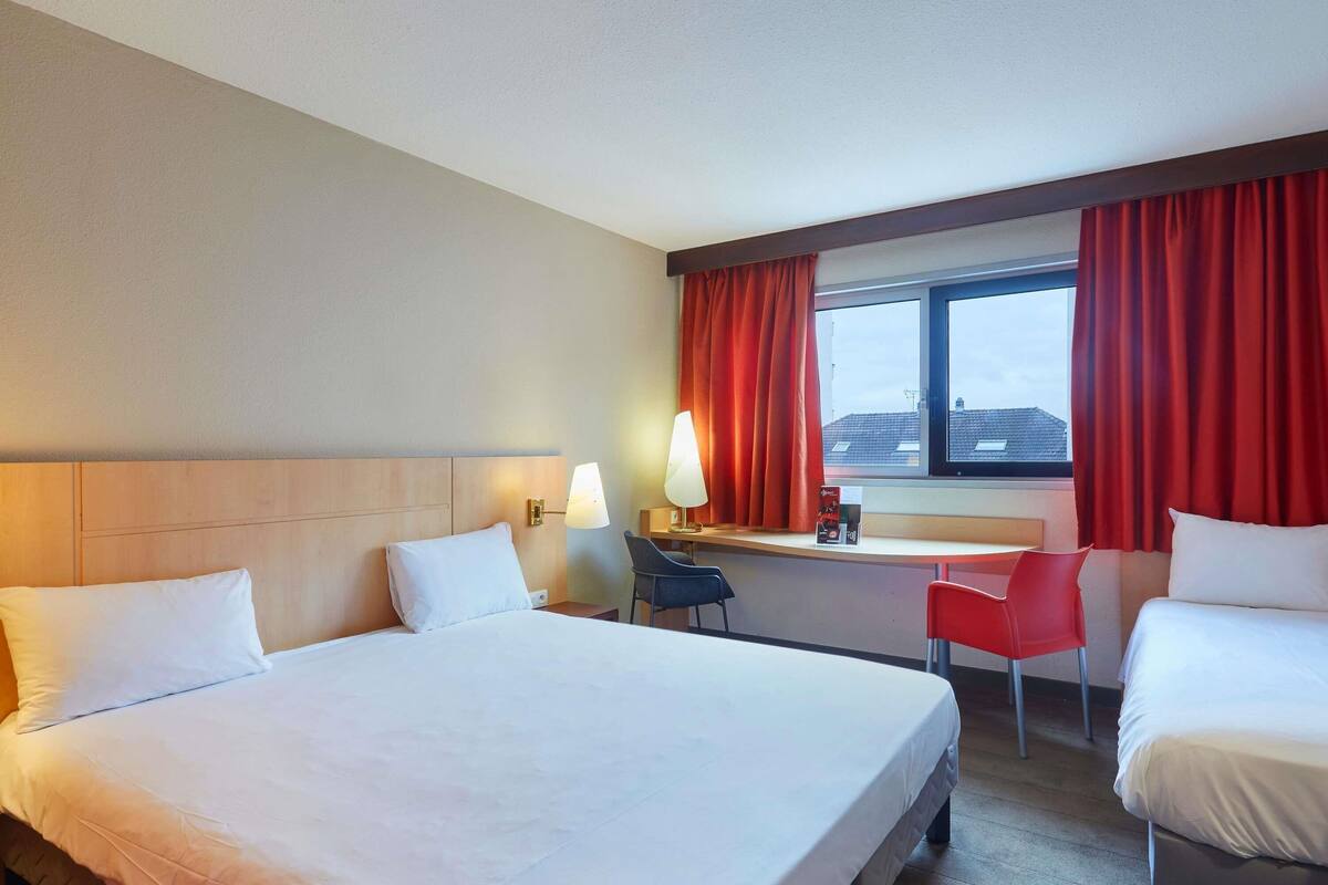 A hotel room with a large bed, a desk with a chair, and a window with red curtains.