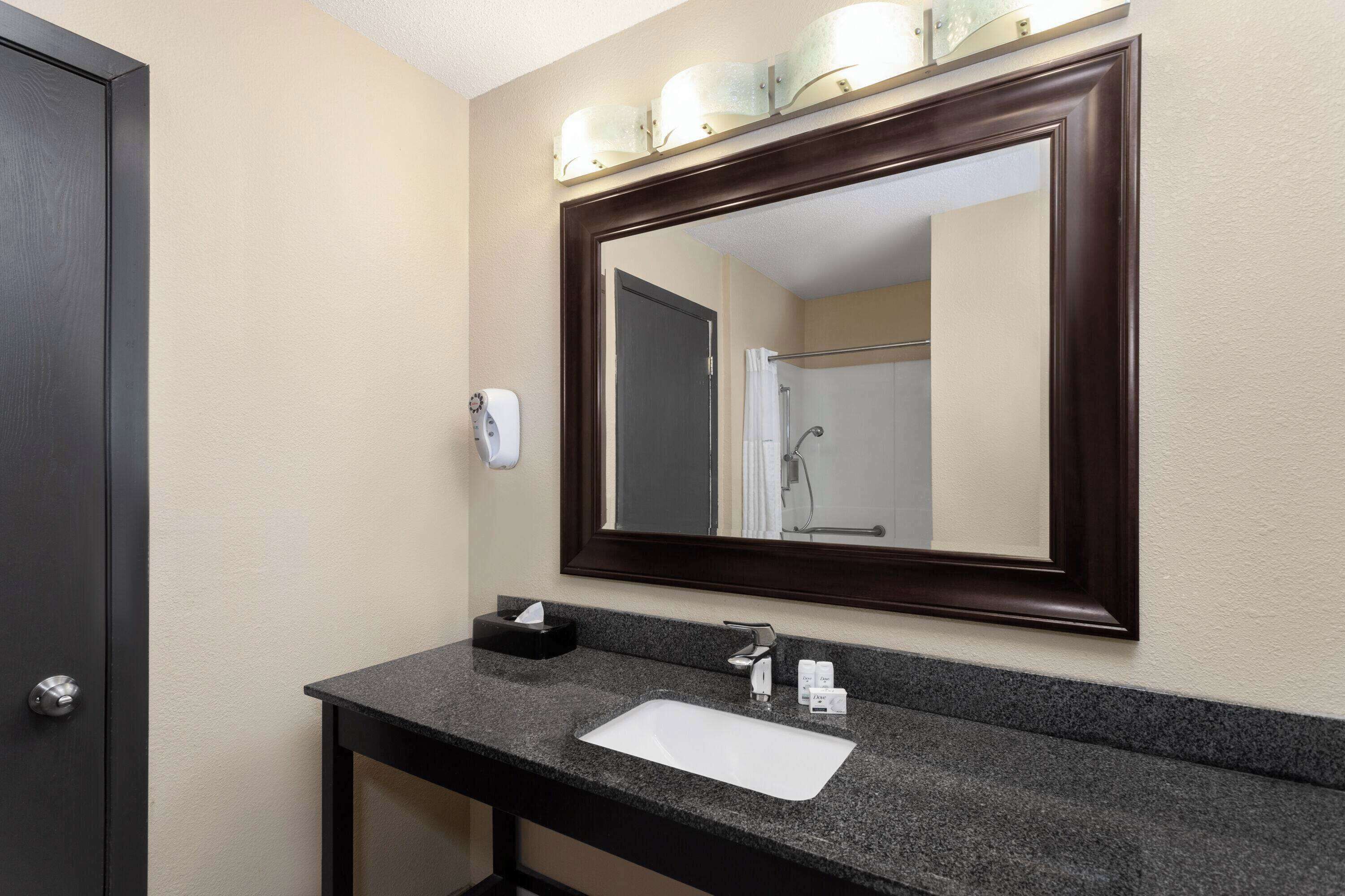 A bathroom with a dark wood-framed mirror, a white sink, a towel dispenser, and a soap dispenser.
