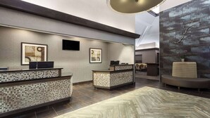 A modern reception area with a herringbone-patterned floor, two reception desks with mosaic tile backs, framed artwork on the walls, and a seating area with a cushioned bench.