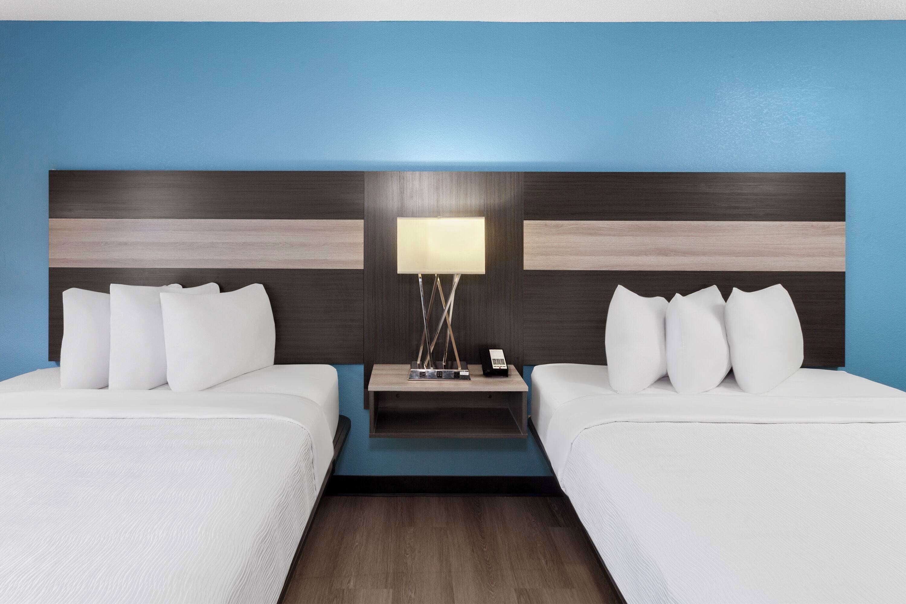 A hotel room with two beds, a nightstand with a lamp, and a blue accent wall.