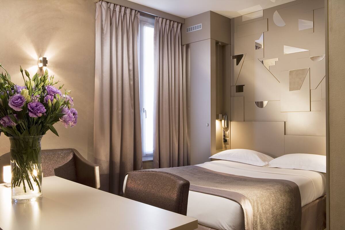 Una habitación de hotel con cama, una mesa con un florero, una silla y una ventana con cortinas.