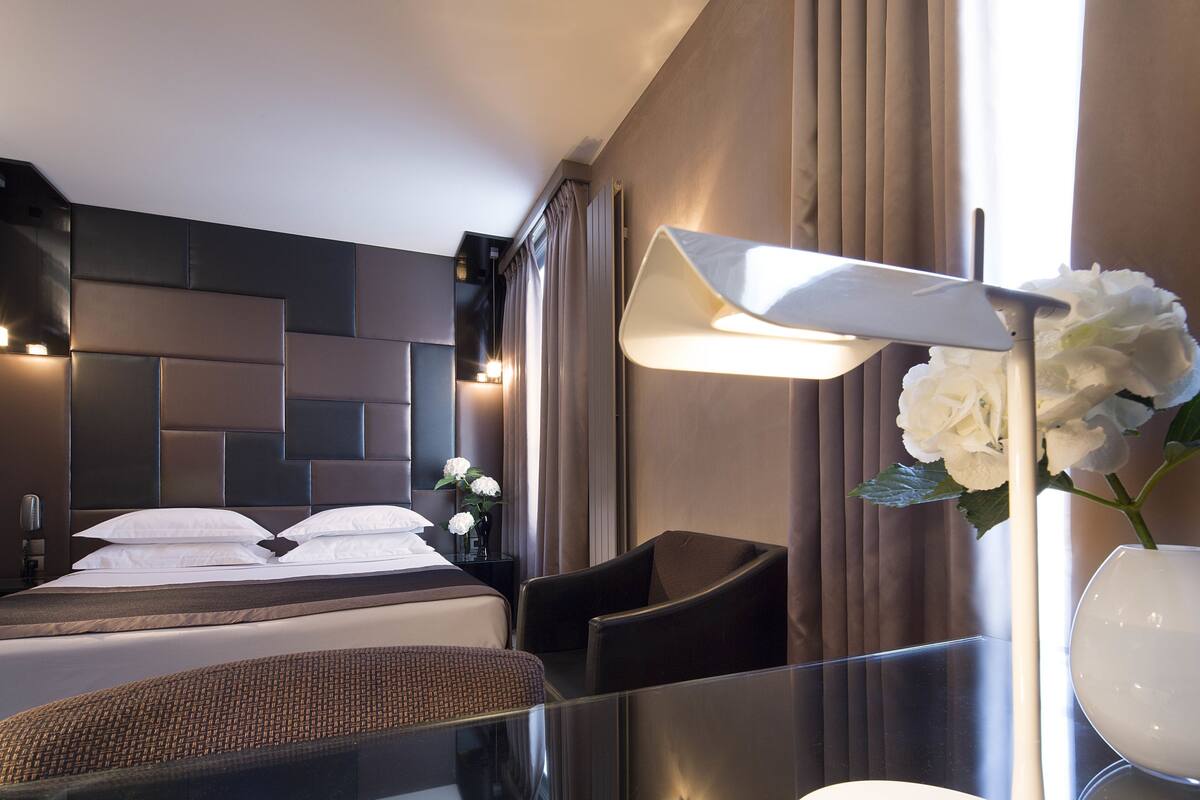 Una habitación de hotel moderna con una cama grande, un escritorio, una silla, una lámpara y un florero con flores.