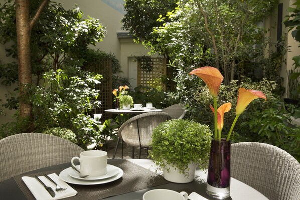 An outdoor dining area with wicker chairs, a table set with white plates, cutlery, and a white cup. There are potted plants and a vase with flowers on the table. - Hôtel A La Villa des Artistes (Paris)