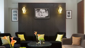 Un espacio lounge moderno con sofás negros, cojines amarillos y verdes, una mesa redonda de vidrio con jarrones de flores y cuadros en las paredes.