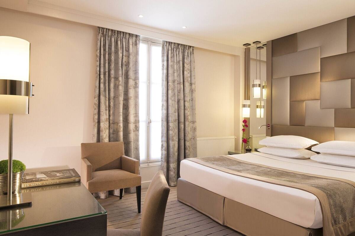 Una habitación de hotel con cama, una silla, un escritorio con lámpara y una ventana con cortinas.