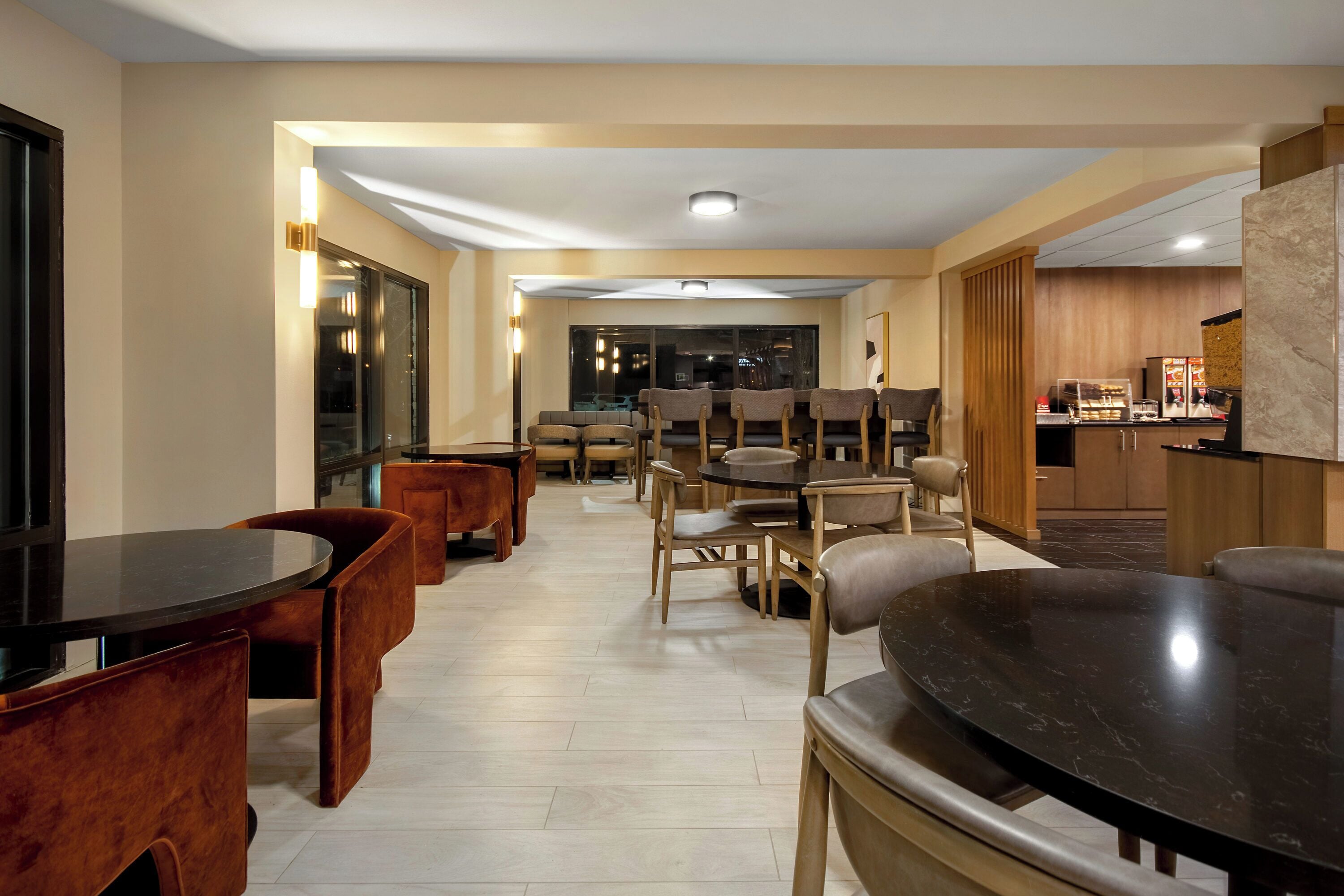 A modern dining area with multiple tables and chairs, a kitchen in the background, and a view of the city at night.