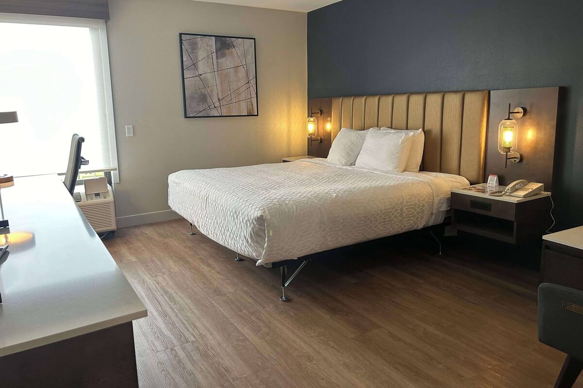 A hotel room with a bed, bedside table, lamp, and a desk with a chair.
