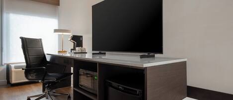 A modern office space with a large flat-screen TV, a black office chair, a desk with a lamp, a microwave, and a dishwasher.