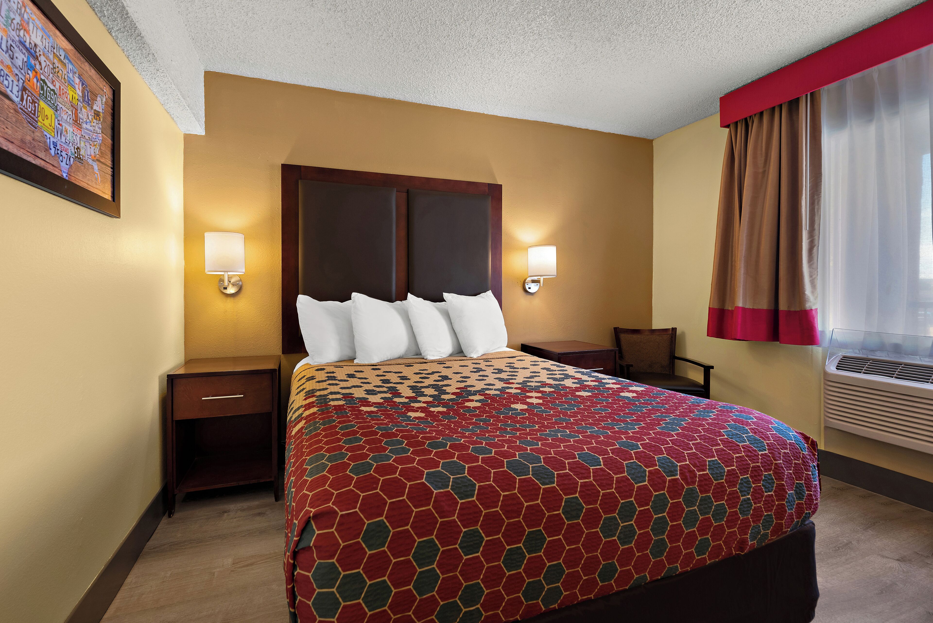 A hotel room with a large bed, two bedside tables, a nightstand, a wall-mounted lamp, and a window with curtains.