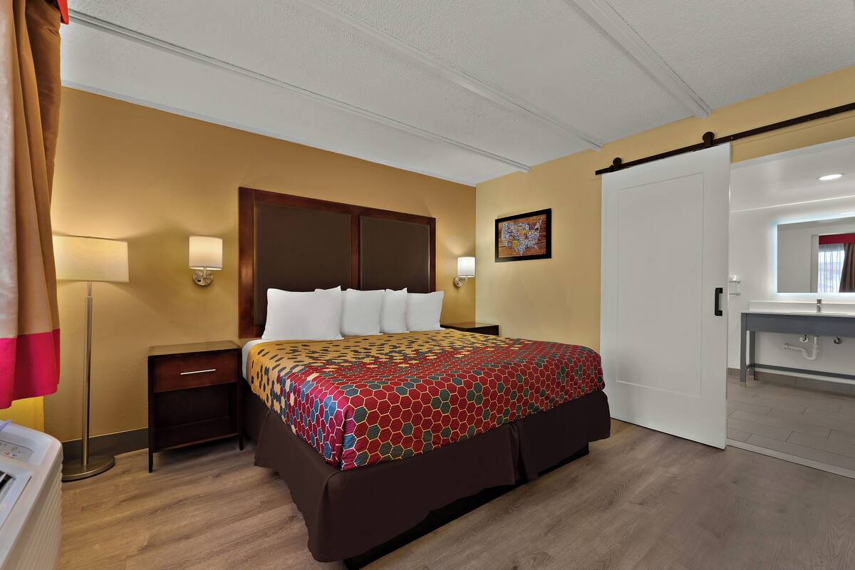 A hotel room with a bed, a nightstand, a lamp, a painting, and a bathroom door.