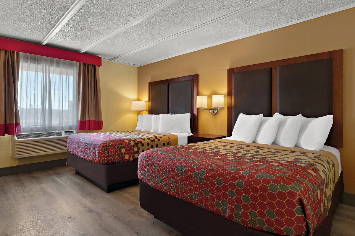 A hotel room with two beds, a window with curtains, wall-mounted lamps, and an air conditioning unit.