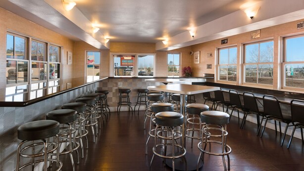 A spacious bar area with multiple stools and a long counter, large windows along the walls, and a visible exit door. - Kings House Motor Hotel, a Dazzler Select by Wyndham (Flagstaff)