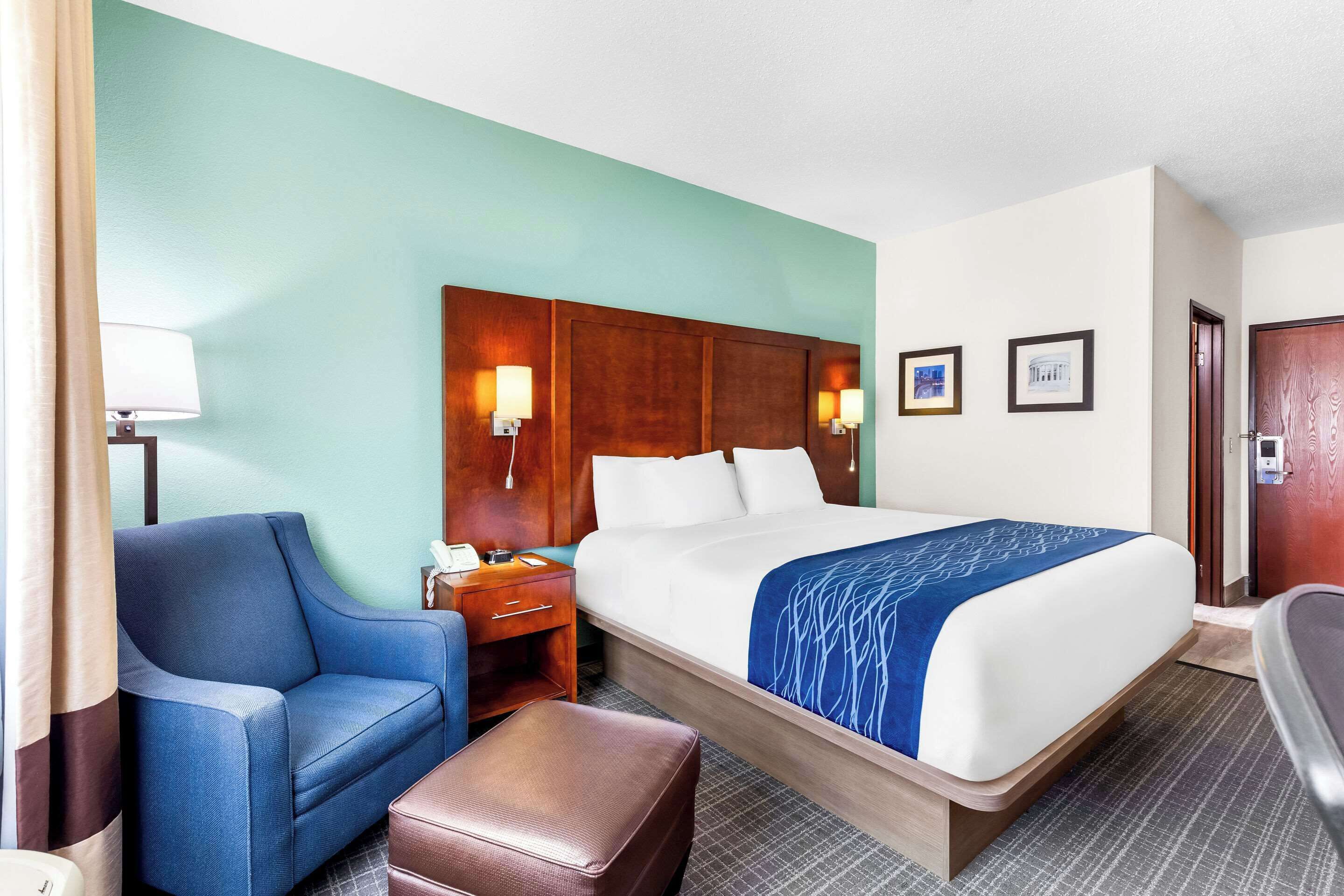 Una habitación de hotel con una cama, un sillón azul, una mesita de noche de madera, un reposapiés marrón y una pared decorada con cuadros.