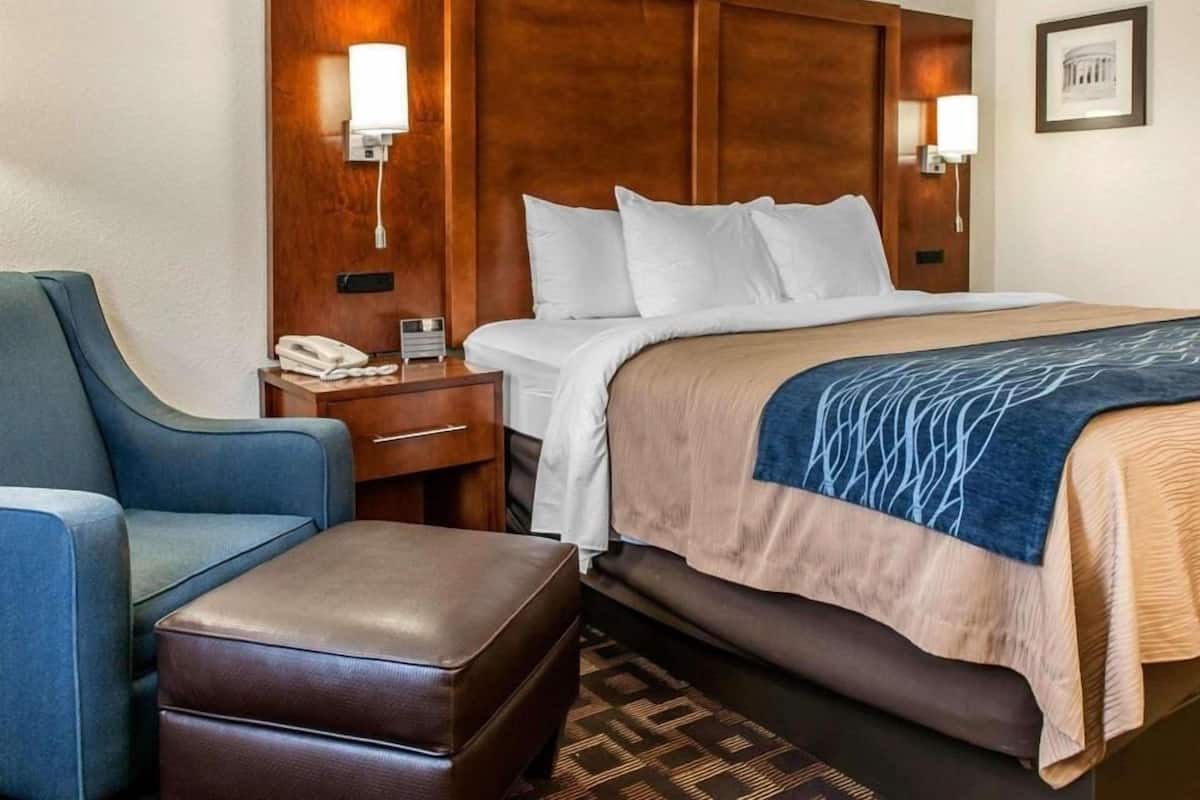 A hotel room with a bed, a desk with a phone, a chair, and a small ottoman.
