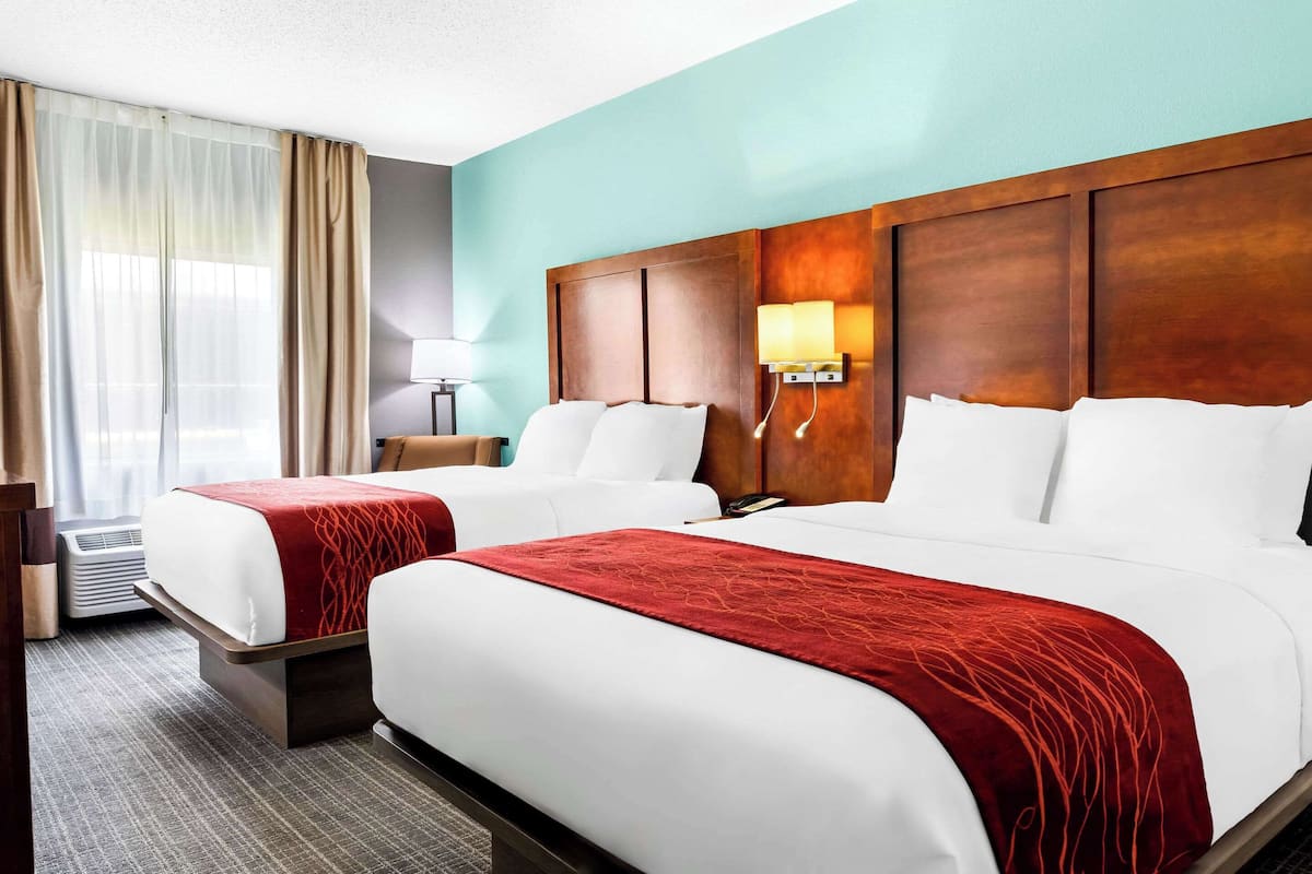 A hotel room with two beds, a wooden headboard, a red accent pillow, a lamp, and a window with curtains.