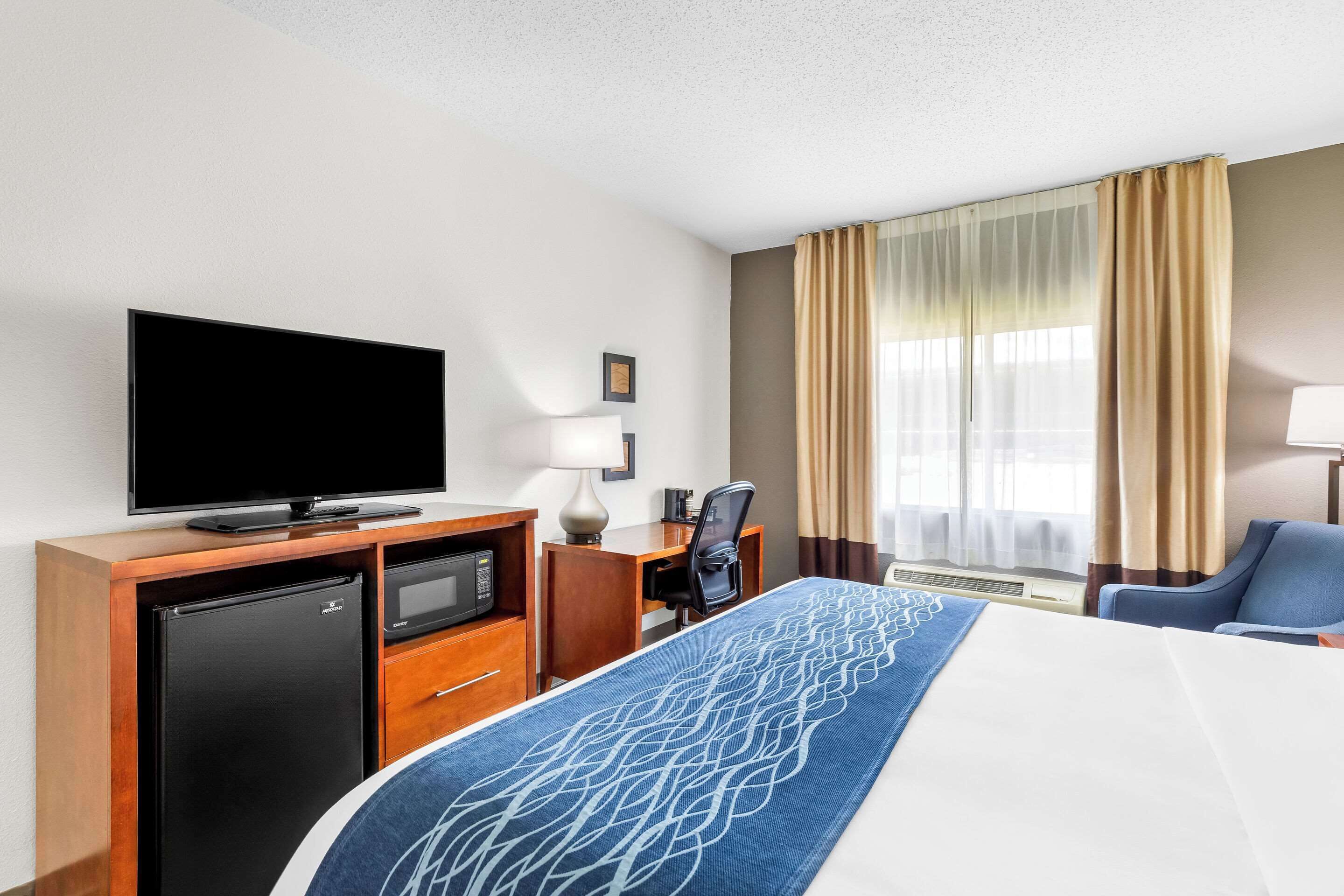 A hotel room with a bed, a desk with a computer, a TV, a mini-fridge, and a window with curtains.