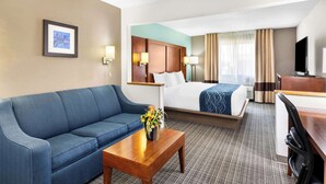A hotel room with a blue sofa, a wooden coffee table with a flower vase, a bed with a blue blanket, a desk with a television, and a framed picture on the wall.